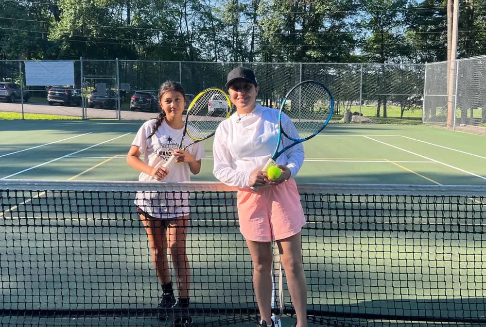 A junior player and an adult coach stand together at the net holding racquets and a tennis ball.